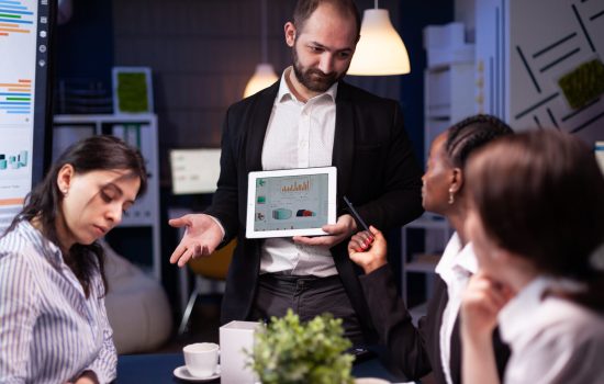 Focused workaholic entrepreneur man working overtime presenting company statistics Focused workaholic entrepreneur man working overtime presenting company statistics using tablet. Diverse multi-ethnic businesspeople overworking in office meeting room late at night.